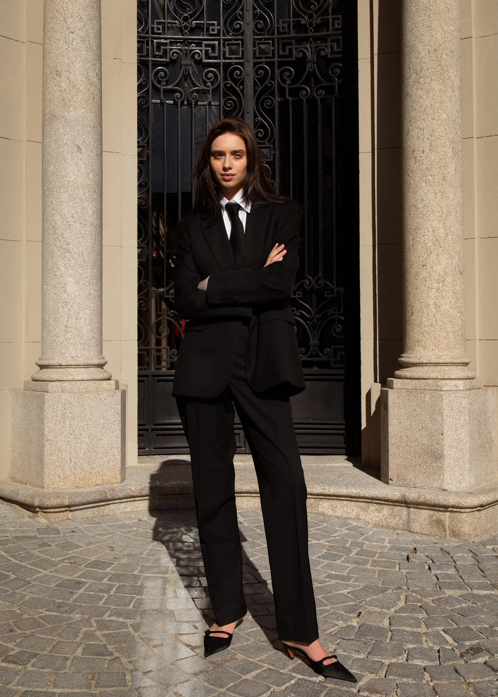 Maison Sarava women's black single belted blazer and trousers suit with tie. Model stands with arms crossed.