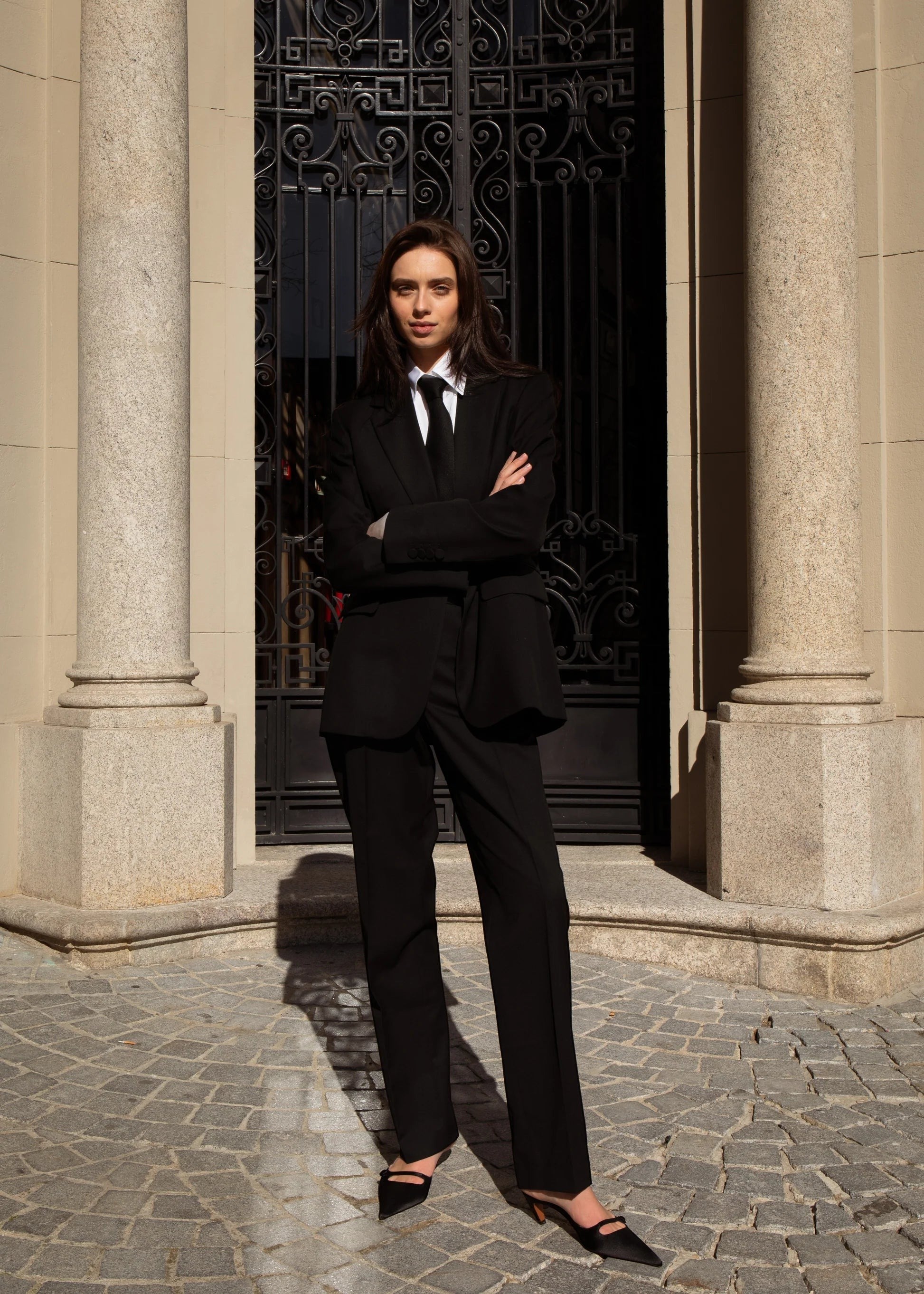 Maison Sarava women's black single belted blazer and trousers suit with tie. Model stands with arms crossed.