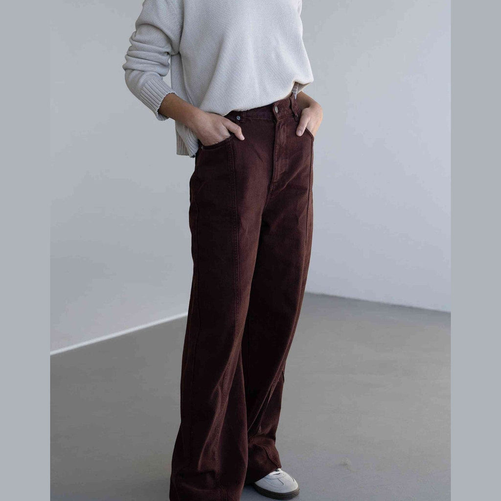 Maison Sarava brown denim trousers with a high rise waist and wide leg silhouette, worn by a model with hands in pockets.