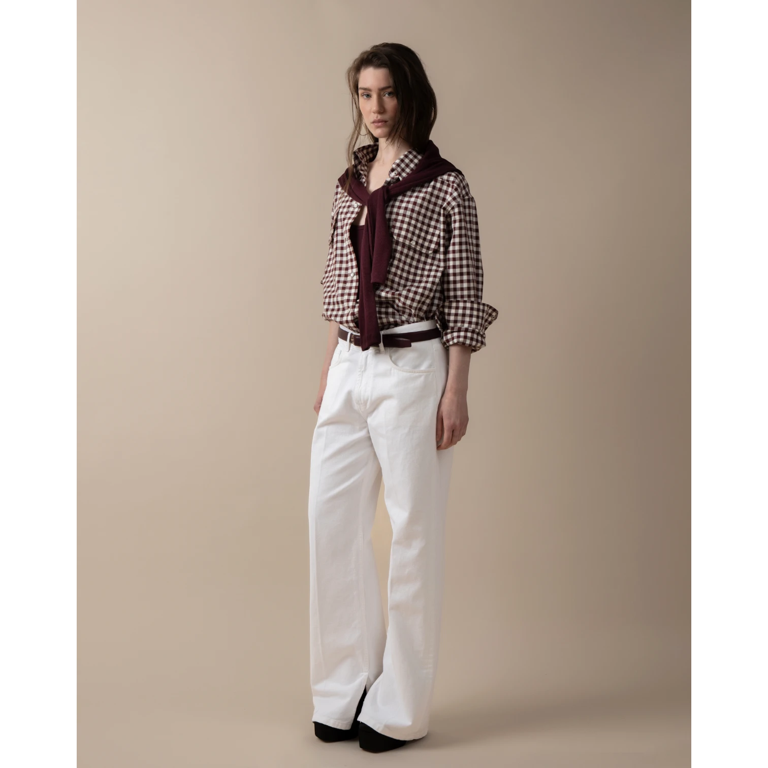 Maison Sarava women's oversized brown and white gingham western shirt with pointed collar, buttoned front, and patch pockets. Model wears white wide-leg pants and a burgundy sweater.